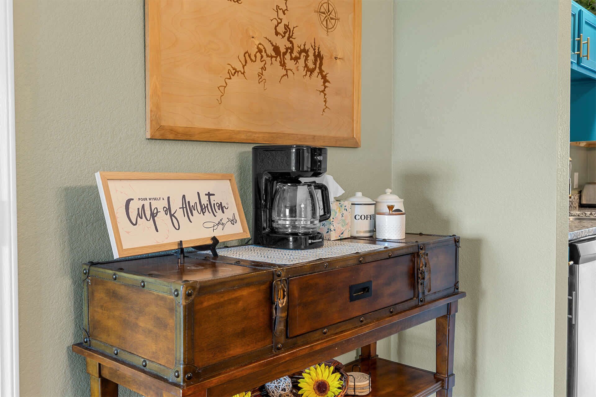 Un mobiletto in legno dall'aspetto antico, con una macchina per il caffè, un'immagine incorniciata con una citazione motivazionale e una decorazione a girasole.