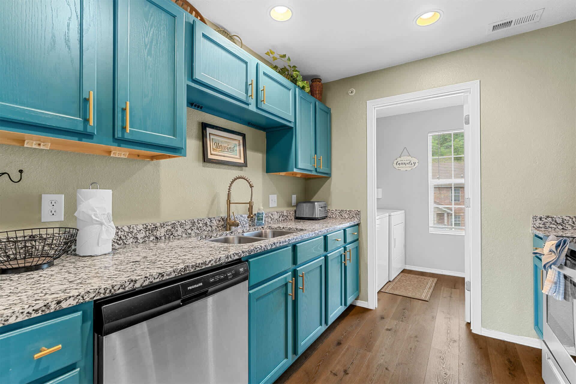 A kitchen with teal cabinets, a granite countertop, a dishwasher, and a window with blinds.