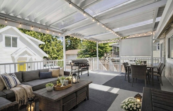 Een overdekt terras met een barbecue, buitenmeubilair en uitzicht op een woning.