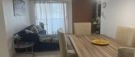 A dining area with a wooden table, chairs, a small side table with a vase, and a sofa with cushions.