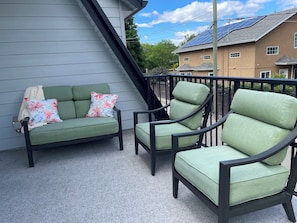 Uma varanda com sofá e poltronas verdes, guarda-corpo e vista para uma casa com painéis solares.