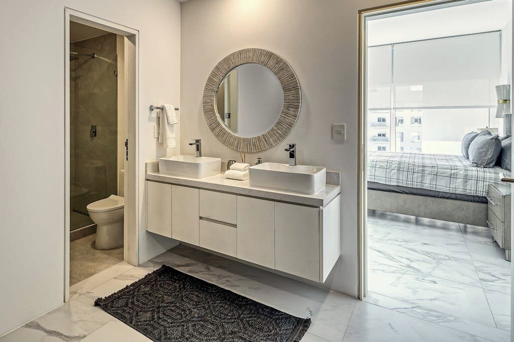 A modern bathroom with a walk-in shower, two sinks, a large mirror, and a view of a bedroom with a bed and a window.