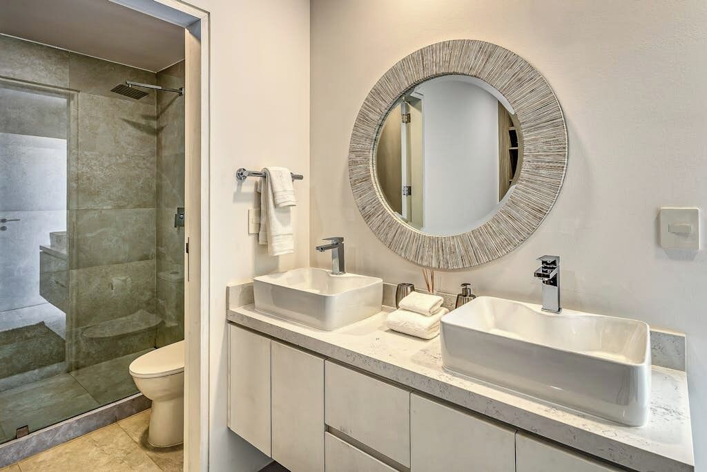 Een badkamer met twee wastafels, een ronde spiegel en een douchecabine van glas.
