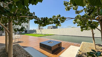 Un espacio exterior moderno con terraza de madera, un pozo de fuego cuadrado y una piscina.