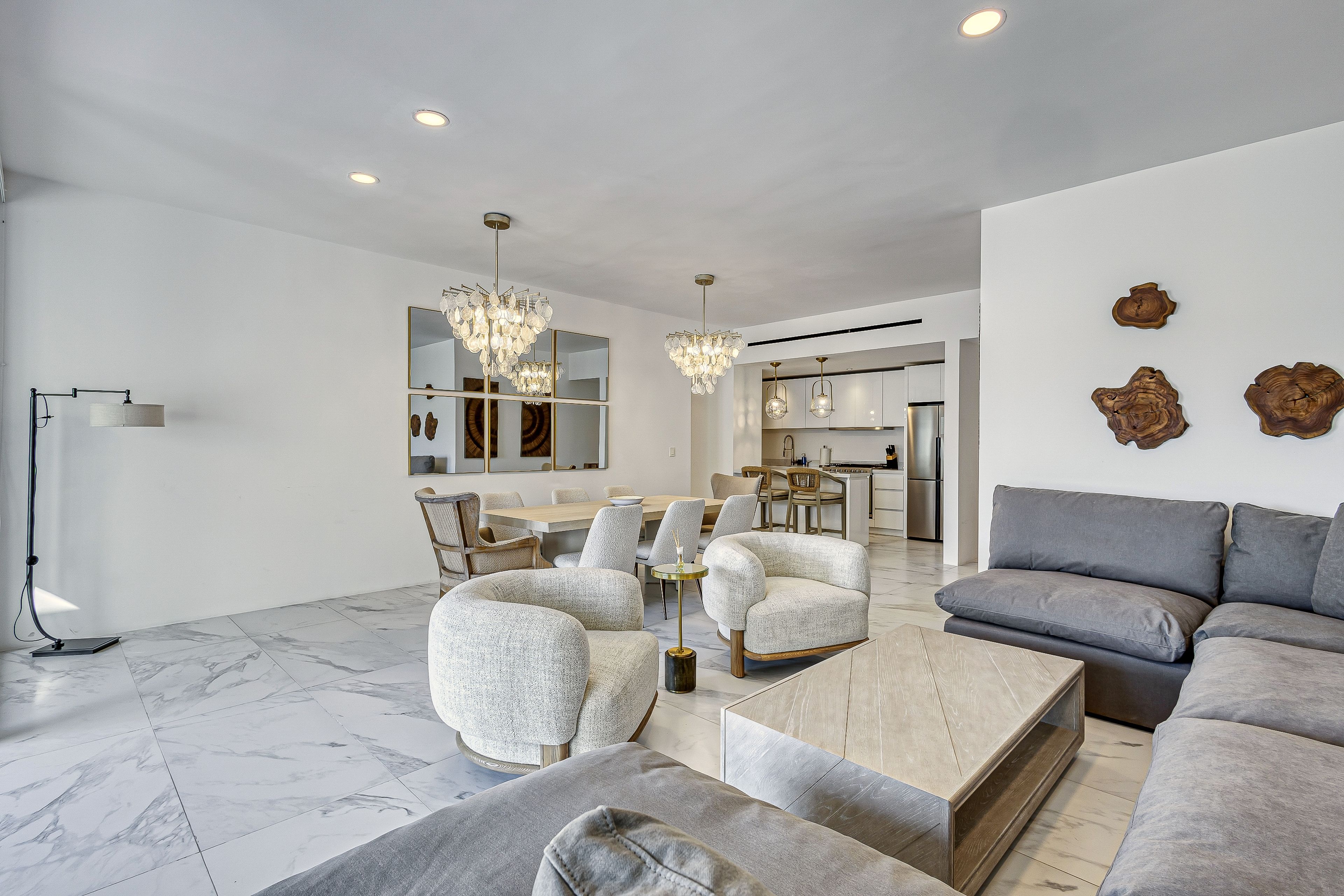 A modern living room with a gray sofa, armchairs, and a coffee table. There is a dining area with a table and chairs, and a kitchen in the background.