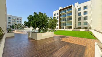 Un espacio exterior moderno con terraza de madera, jardineras con árboles y césped bien cuidado.