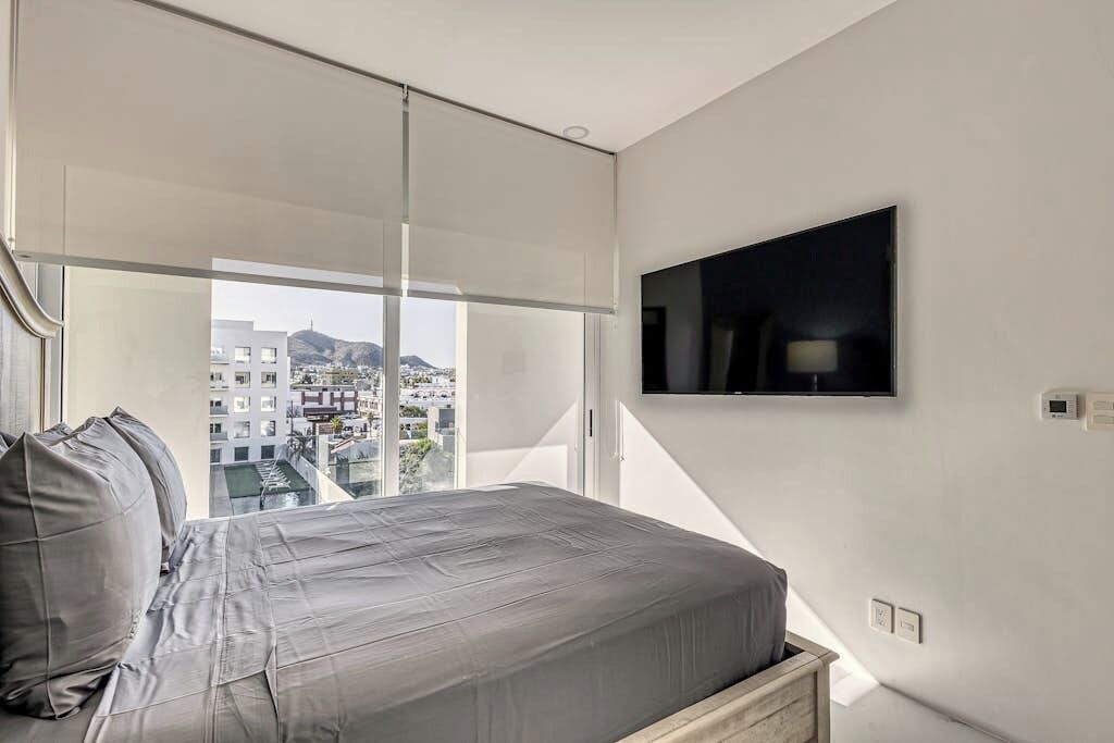 A modern bedroom with a bed, a TV mounted on the wall, and a large window offering a city view.