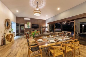 A modern dining area with a wooden table set for six, a large chandelier, and a view of the outdoors. - Casa Pochote | Private Saltwater Pool & Golf Retreat in Hacienda Pinilla (Tamarindo)