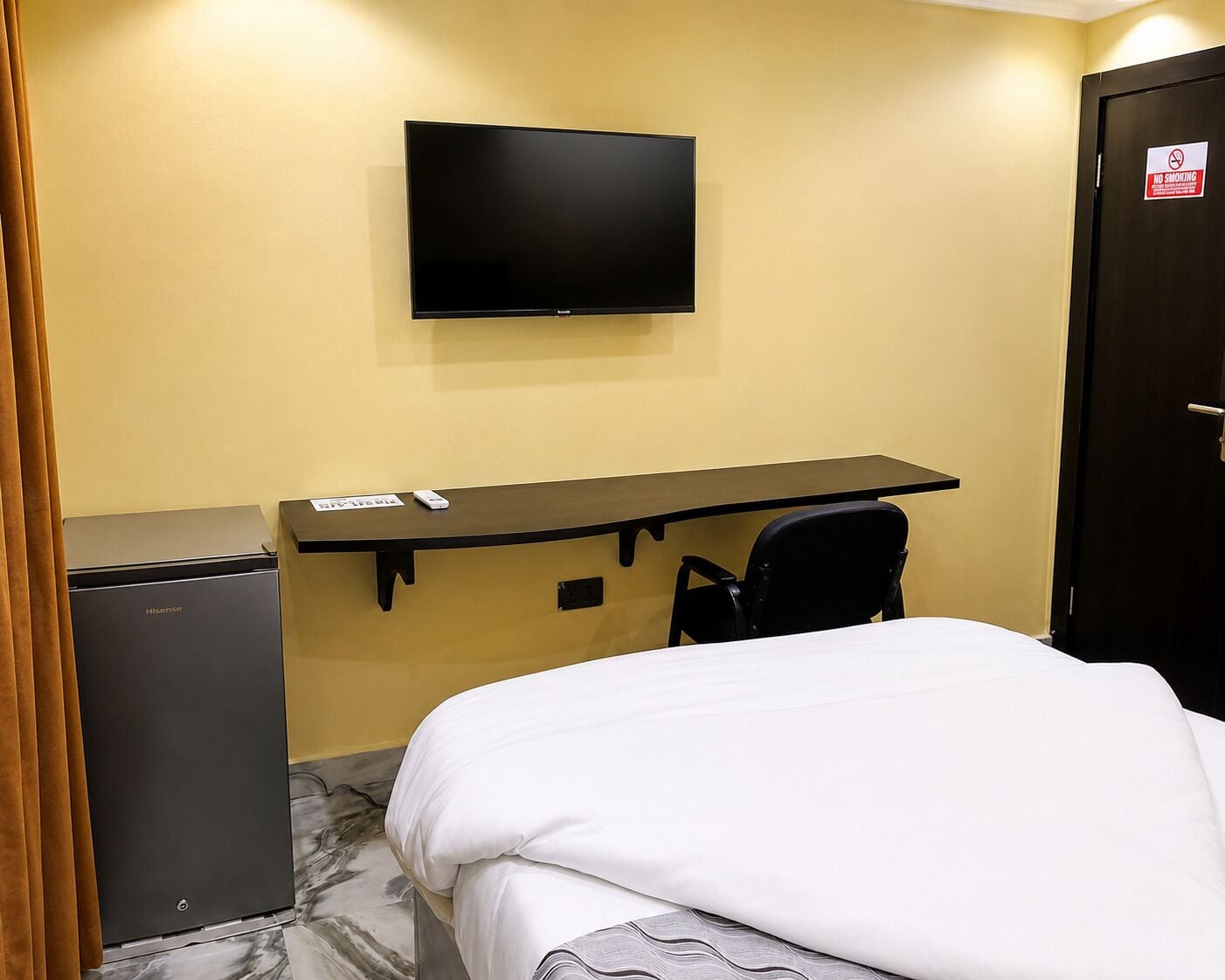 A hotel room with a bed, a desk with a chair, a television mounted on the wall, and a small refrigerator.