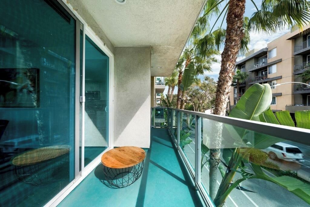 Un balcon avec une porte vitrée, un tabouret en bois et une vue sur des palmiers et un immeuble.