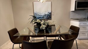 Un comedor con una mesa de vidrio, cuatro sillas, una araña, un cuadro y una cocina al fondo.