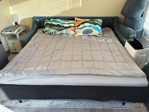 A neatly made bed with a quilted cover, a bedside table with a lamp, and a window with blinds.