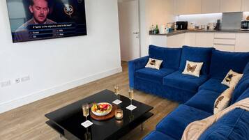 A modern living room with a blue sofa, a black coffee table with a fruit bowl and glasses, and a TV displaying a quiz game.
