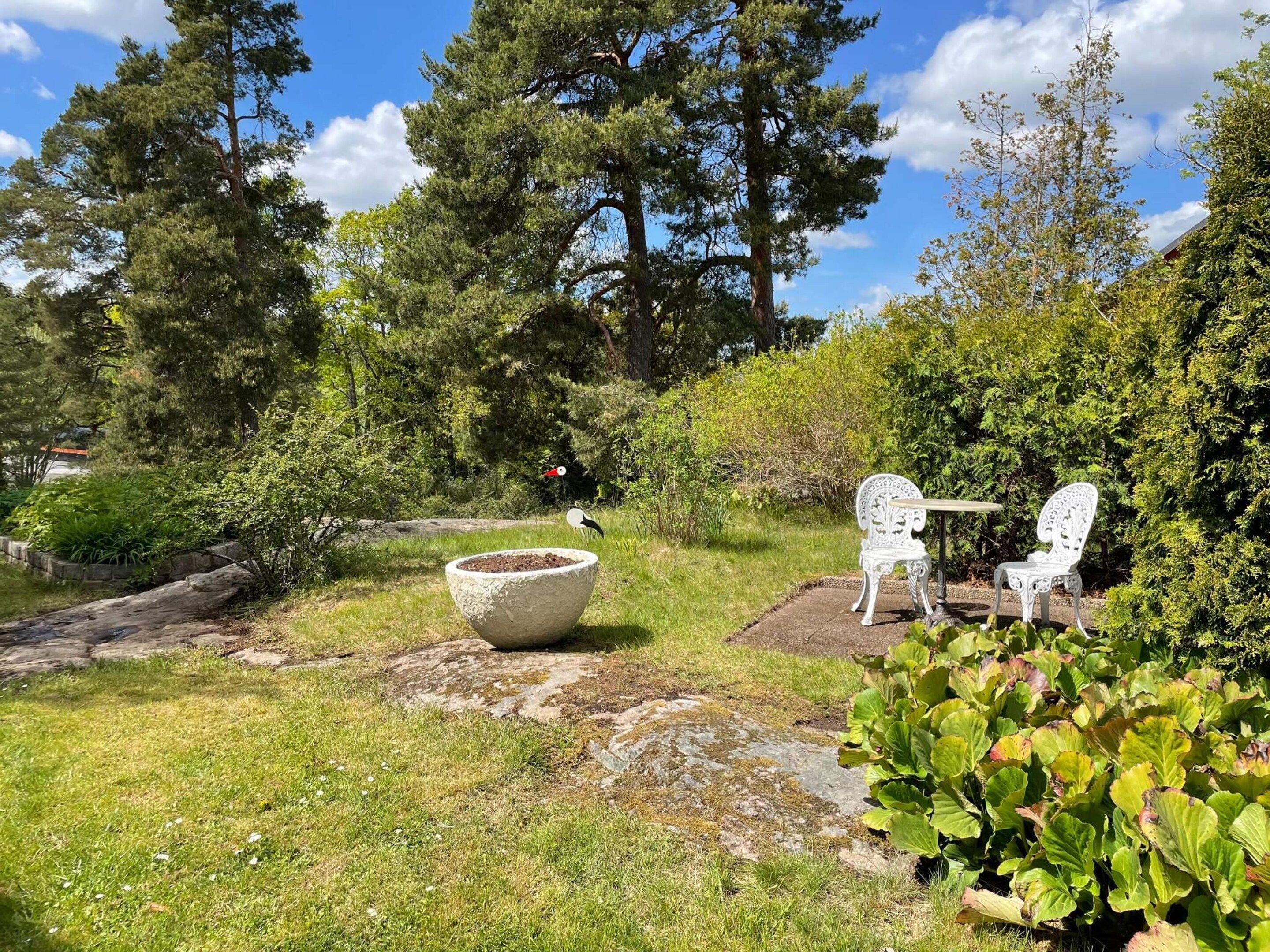 A garden with a stone planter, two white chairs, and a table.