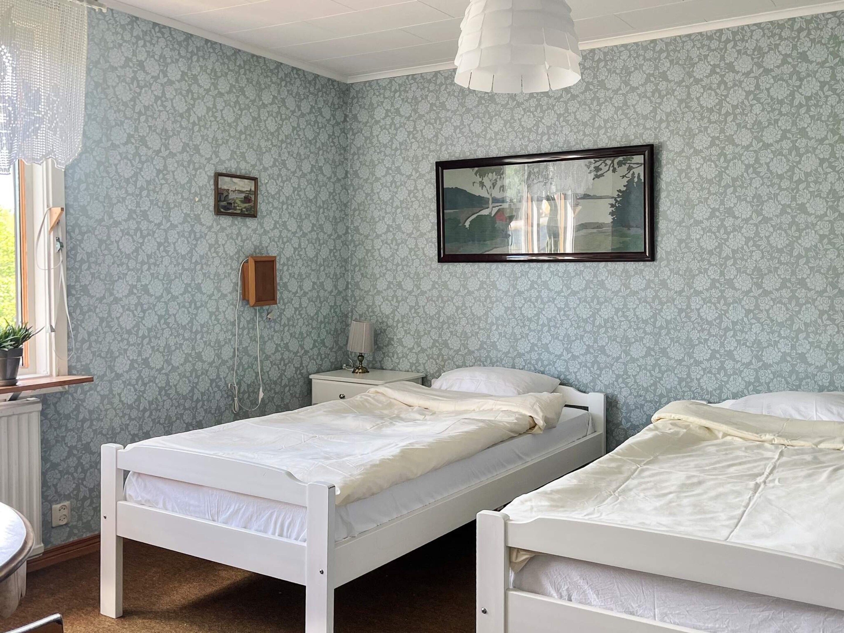 A room with two single beds, a small bedside table with a lamp, a framed picture on the wall, and a window with a curtain.