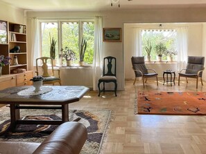 A living room with a wooden floor, a brown leather sofa, a coffee table, two armchairs, a bookshelf, and a window with plants. - 5 Person Holiday Home in Gunnebo (Gunnebo)
