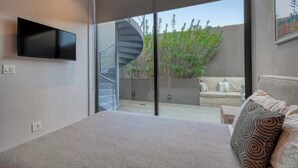 Ein modernes Schlafzimmer mit einem großen Bett, einem Flachbildfernseher, einer Wendeltreppe und Blick auf eine Terrasse mit Gartenmöbeln.