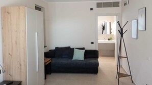 A modern living room with a dark sofa, a wooden wardrobe, a coat rack, and a kitchenette in the background.