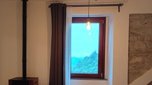 Une pièce avec un poêle à bois, une table avec deux chaises, une fenêtre donnant sur la verdure et un mur en pierre.