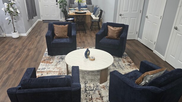 Un salon avec une table basse ronde, quatre fauteuils bleu foncé, un coin repas avec une table dressée pour deux personnes et une plante en pot.