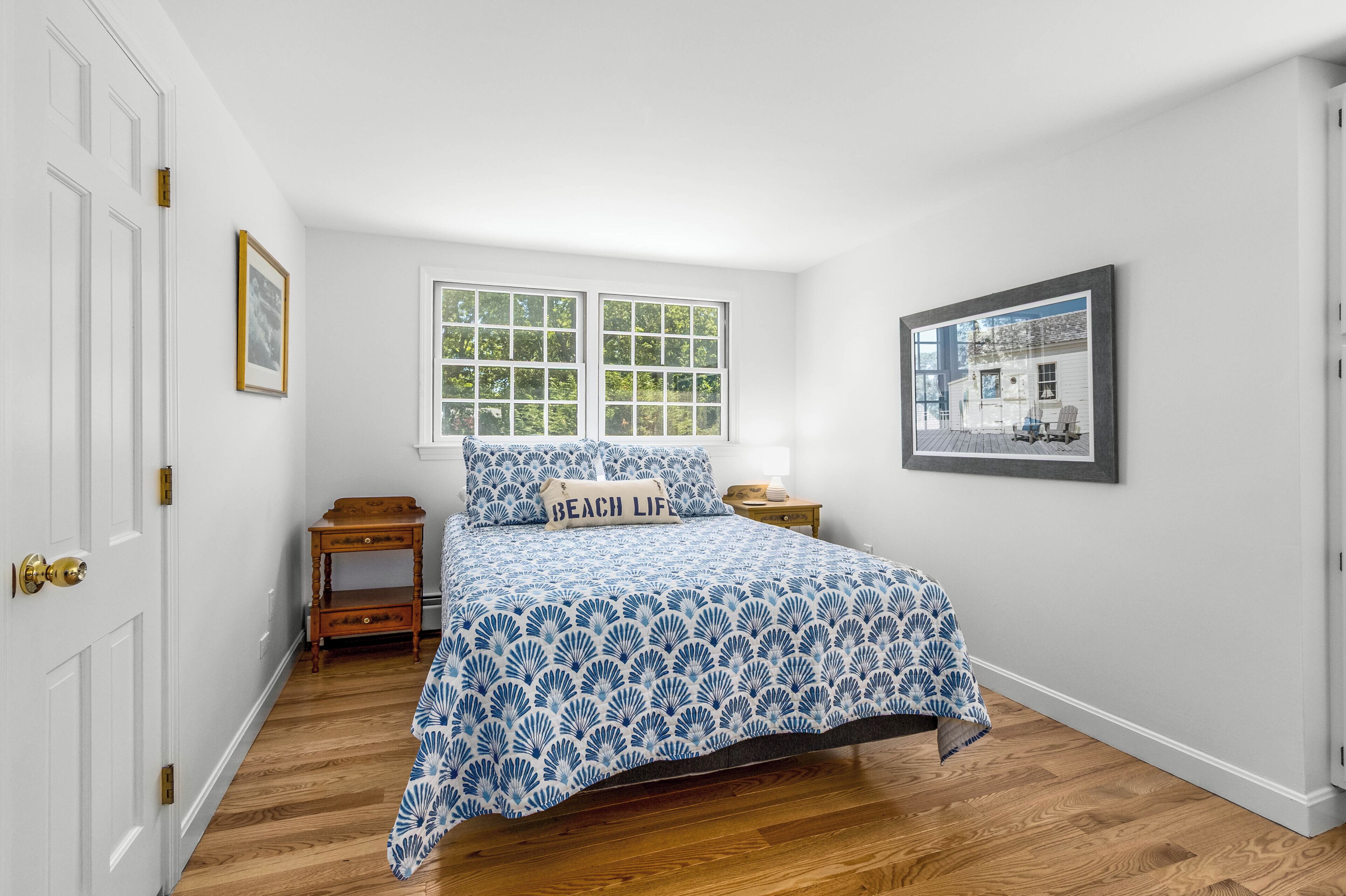 A bedroom with a bed, a window, a nightstand, a framed picture, and a door.