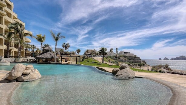 A resort pool with clear water, surrounded by palm trees and a view of the sea. - Stylish Coastal Retreat With Breathtaking Views, Resort Pool & Private Balcony (Cabo San Lucas)