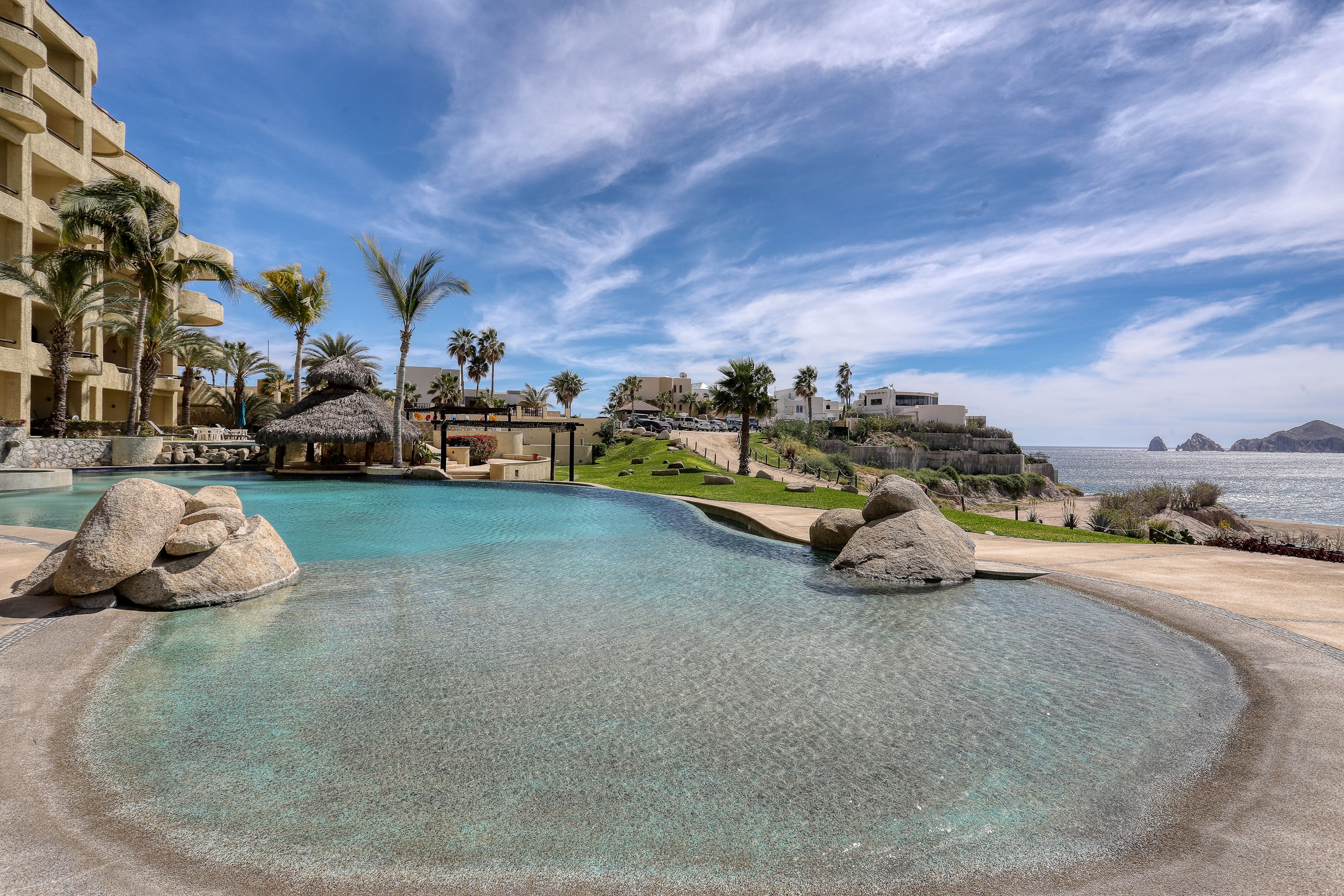 A resort pool with clear water, surrounded by palm trees and a view of the sea.