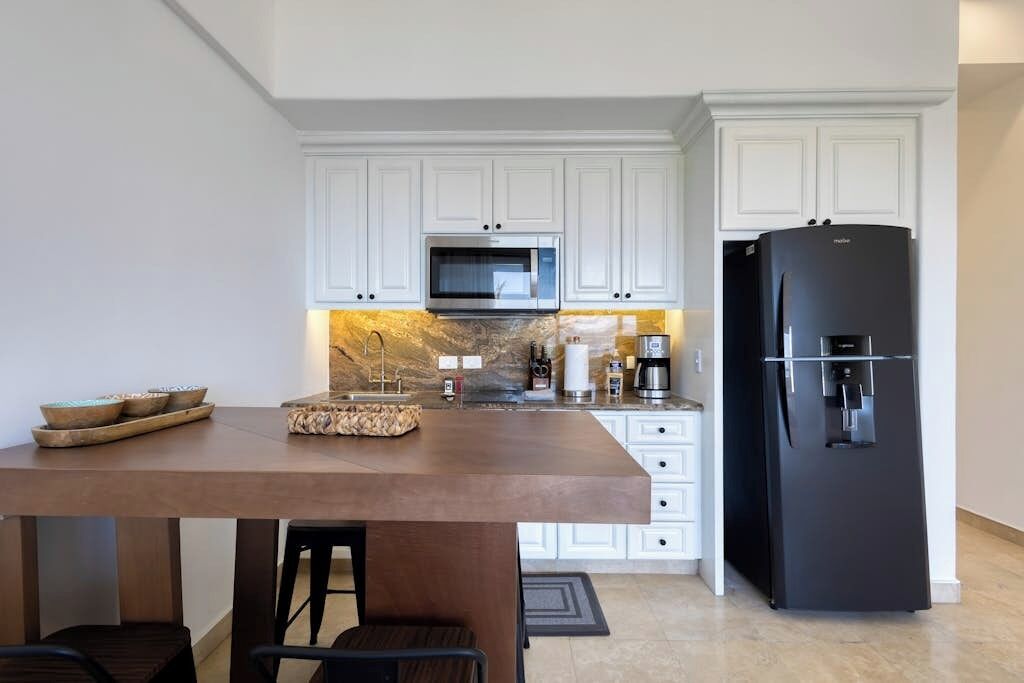 A modern kitchen with white cabinets, a stainless steel microwave, a black refrigerator, and a wooden dining table.