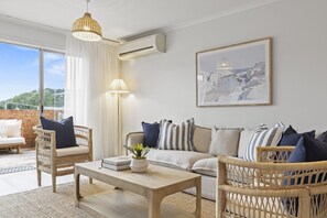 A modern living room with wicker furniture, a wooden coffee table, and a large window with a view of greenery. - Casa Munns - In the heart of Merimbula (Merimbula)