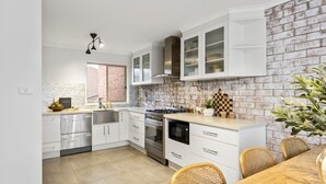 Una cocina moderna con armarios blancos, salpicadero de ladrillo, electrodomésticos de acero inoxidable y una zona de comedor con sillas de mimbre.