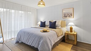 Un dormitorio con cama, lámparas de noche, un cuadro en la pared y una ventana con cortinas de gasa.