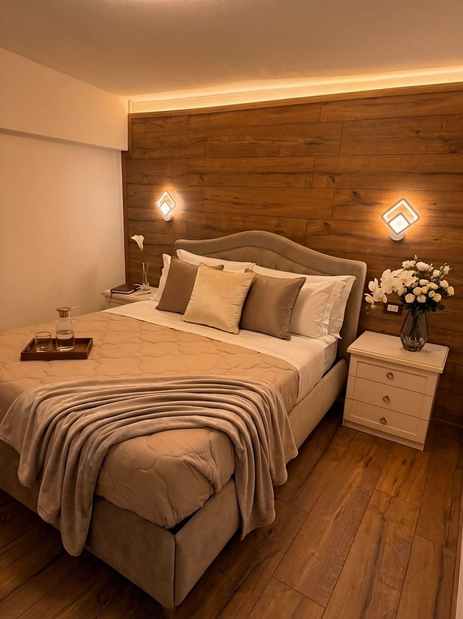 A bedroom with a wooden headboard, a bed with beige bedding, bedside tables with lamps, and a nightstand with a vase of flowers.