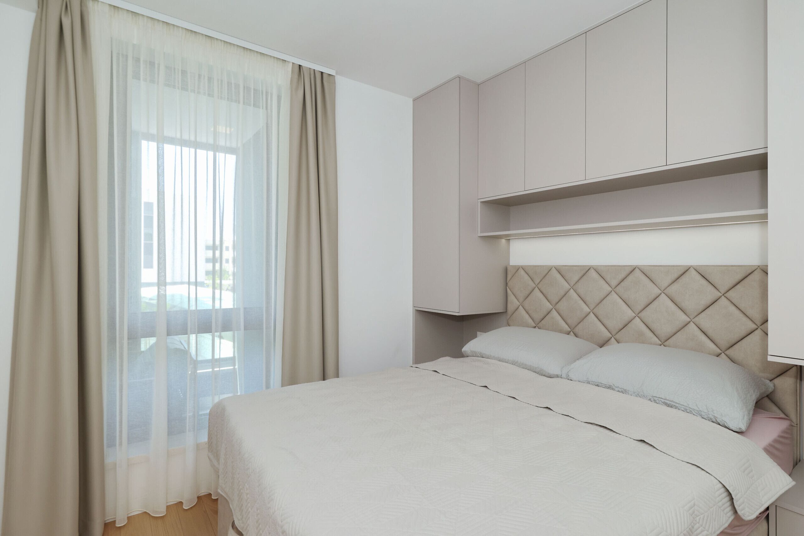 A bedroom with a bed, a window with sheer curtains, and built-in shelving above the bed.