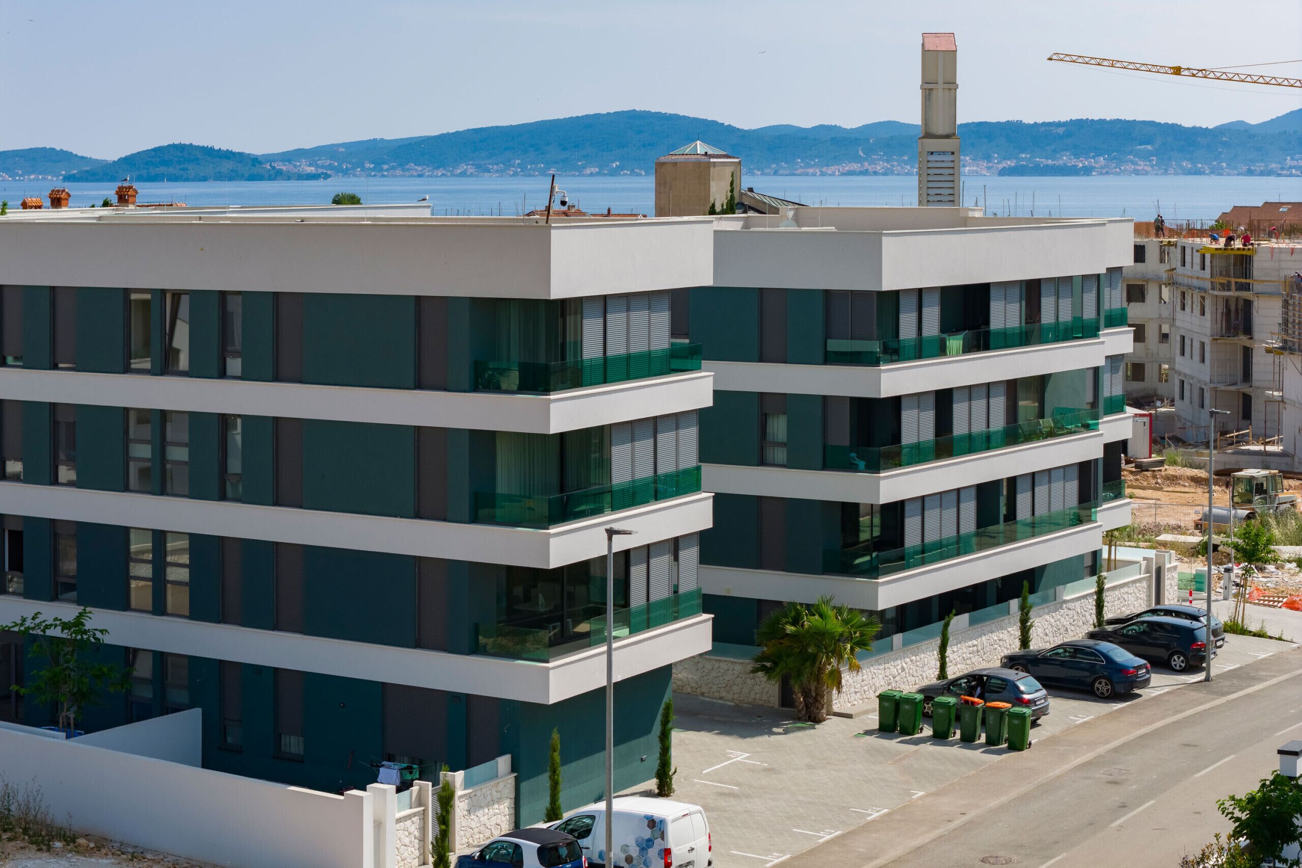 A multi-story building with balconies, a parking area, and a view of the sea and mountains in the background.