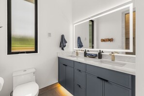 Un baño moderno con inodoro blanco, un espejo grande y un mueble de tocador gris con lavabo.