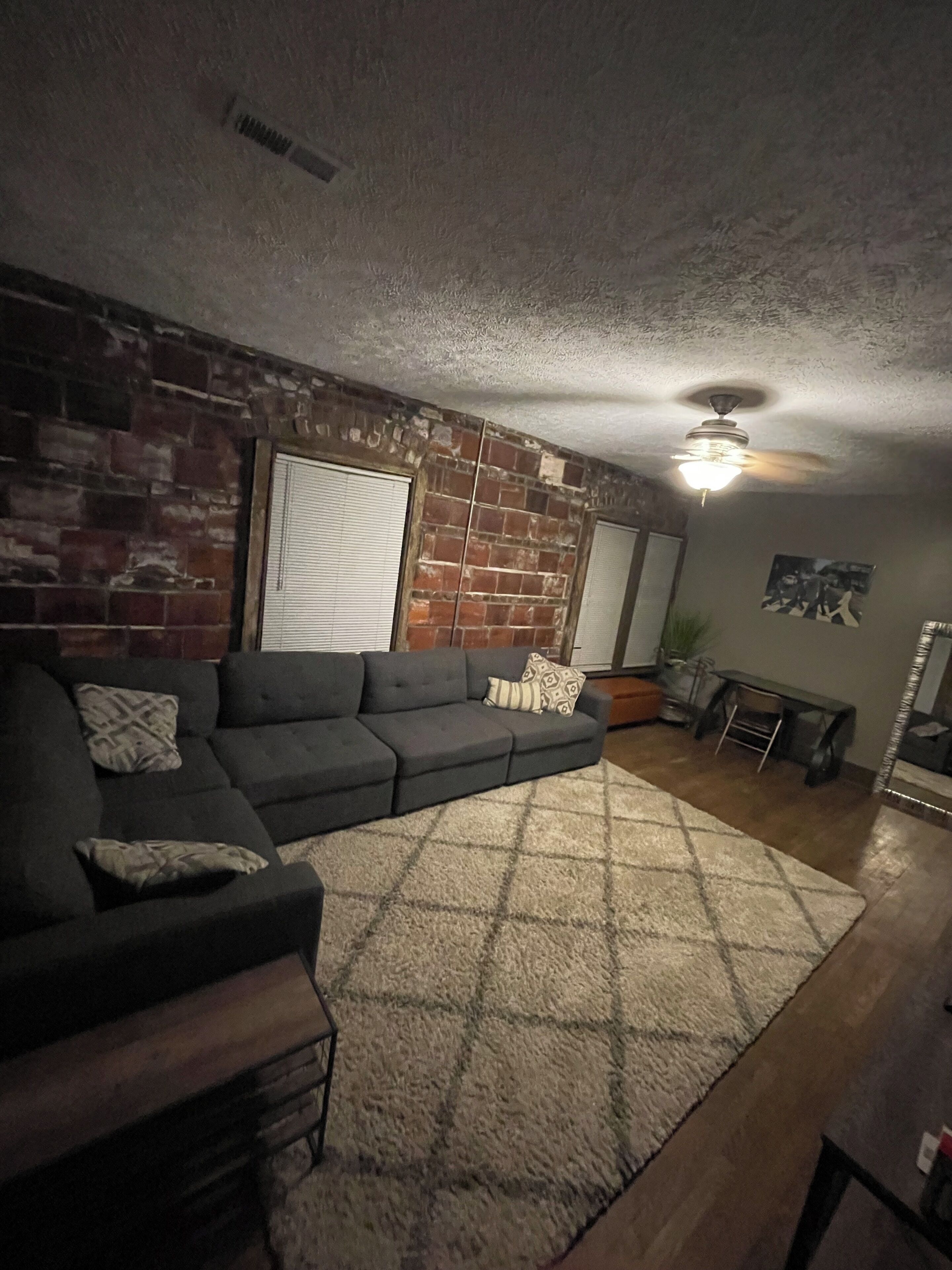 A living room with a sectional sofa, a patterned rug, a ceiling fan, and a brick wall.
