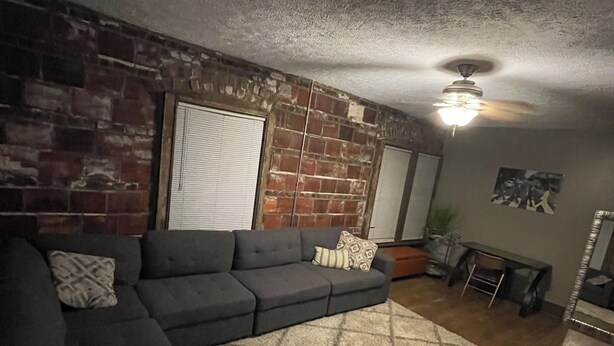Un salon avec un canapé d’angle, un tapis à motifs, un ventilateur de plafond et un mur en briques.