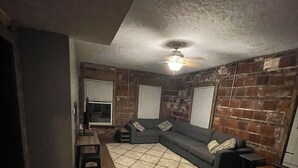 Une pièce avec un plancher en bois, un canapé d’angle, un ventilateur de plafond et des murs en briques.