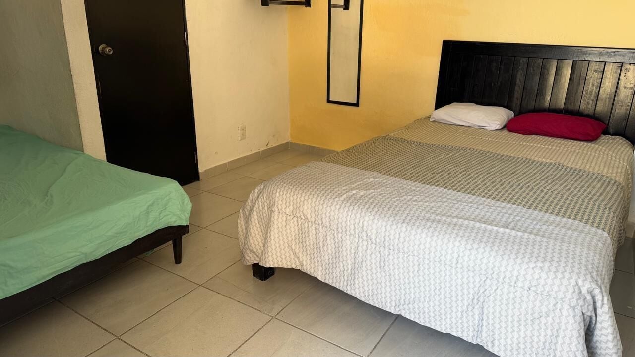 Una habitación con dos camas, una con cubrecama verde y la otra con cubrecama blanca y almohada roja.
