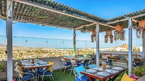 An outdoor dining area with tables and chairs, a pergola with hanging decorations, and a view of a cityscape. - Dog-friendly Loft With Rooftop Infinity Pool, Gym, Balcony & Central AC (Cabo San Lucas)