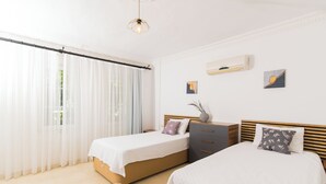 Un dormitori amb dos llits, una finestra amb cortines translúcides, una làmpada al sostre i un aire acondicionat petit fixat a la paret.