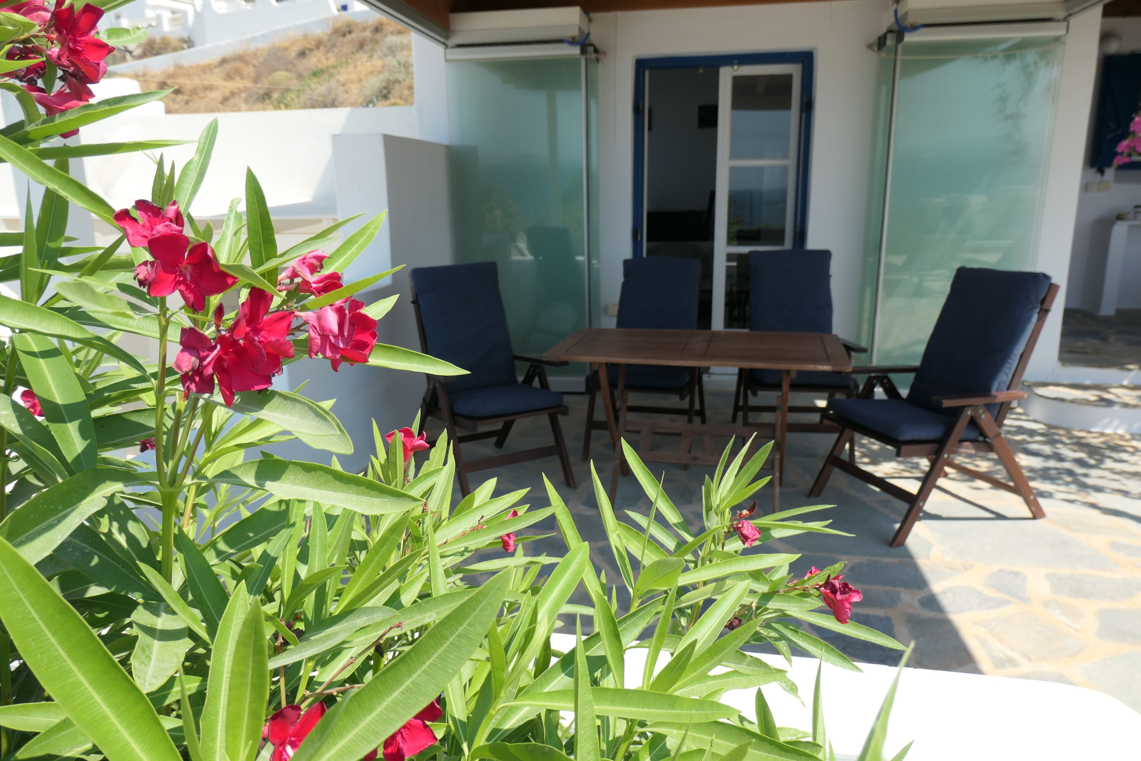 Een terras met een tafel en stoelen, omgeven door groen en een glazen deur naar een kamer.