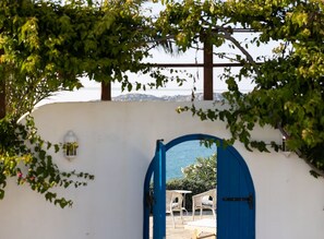 A doorway with a blue arch leading to an outdoor seating area with white chairs and tables, surrounded by greenery and a view of the sea. - Charming Naxos Villa, Ocean Views, Modern Comfort, Tranquil Escape (Naxos)