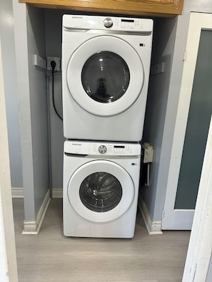 Une machine à laver et un sèche-linge superposés dans une buanderie, avec une étagère en bois au-dessus.