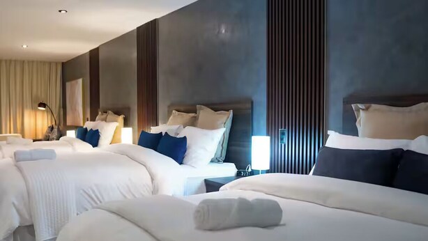 A hotel room with three beds, each with white bedding and multiple pillows. There are bedside lamps and a large window with blinds.