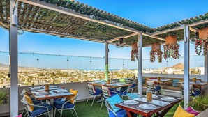 An outdoor dining area with tables and chairs, a pergola with hanging decorations, and a view of a cityscape. - Dog-friendly Apartment With Rooftop Infinity Pool, Gym, Central AC & Balcony (Cabo San Lucas)