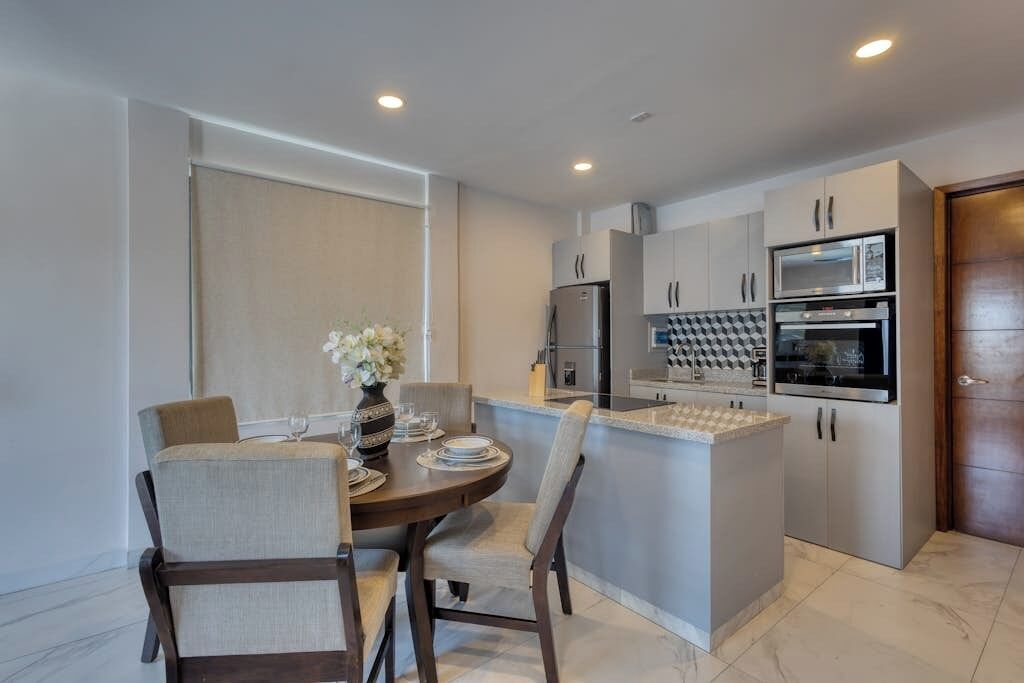 Una cocina moderna con electrodomésticos integrados, un comedor con una mesa redonda y sillas, y un ventanal grande.
