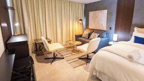A hotel room with a bed, two chairs, a small table, a sofa, a desk, and a large window with curtains.