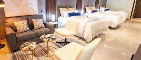 A modern hotel room with a sofa, two armchairs, a small table, a large bed with multiple pillows, and a large abstract painting on the wall.