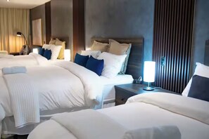 A hotel room with three beds, a bedside table with a lamp, and a large window with curtains.